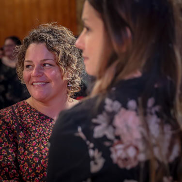 Smiling woman with curly hair in a floral dress looks to the side during a social gathering.