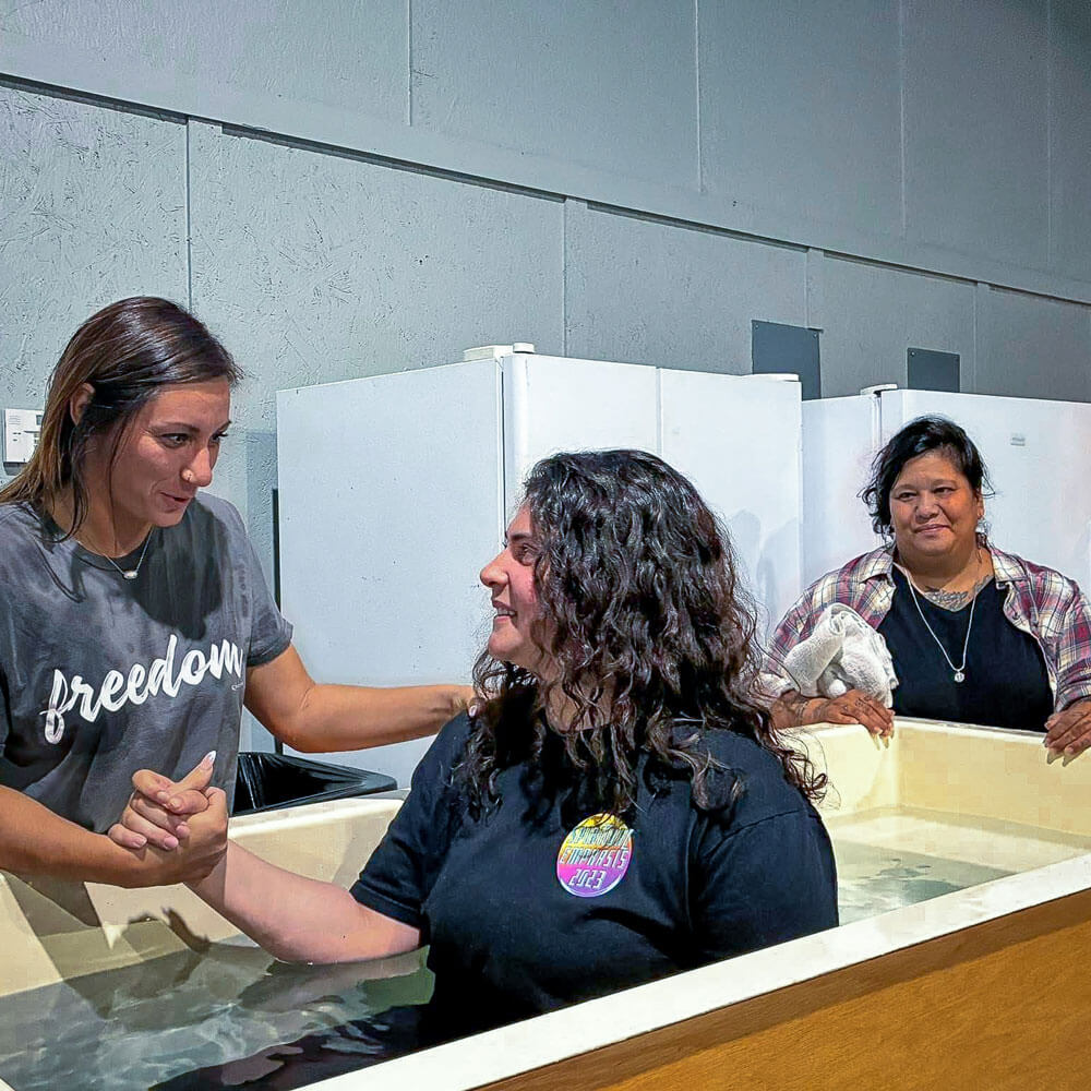 Woman in a freedom t-shirt supports another woman being baptized in a tub, while a third woman observes with a smile.
