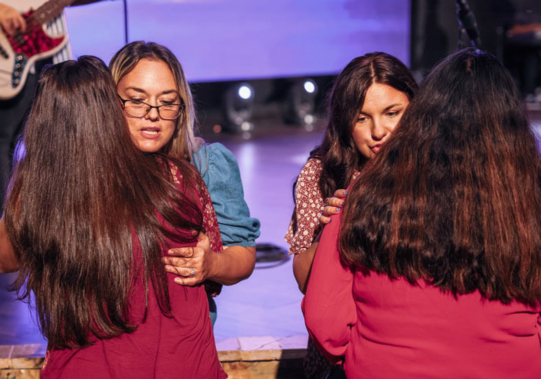 Two pairs of women standing face to face share emotional moments in a setting with stage lighting in the background.
