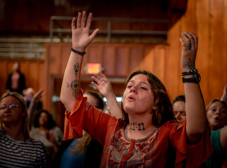 Young woman with tattoos raises her hands in worship during a church service, surrounded by others in prayer.
