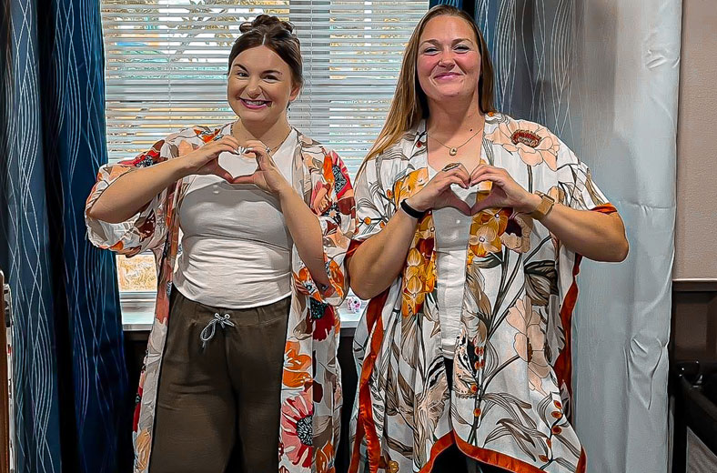 Two smiling women wearing matching floral robes form heart shapes with their hands in front of a sunlit window.
