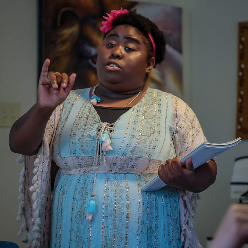 Woman with a flower in her hair speaks passionately while holding a notebook, gesturing with her hand during a presentation.
